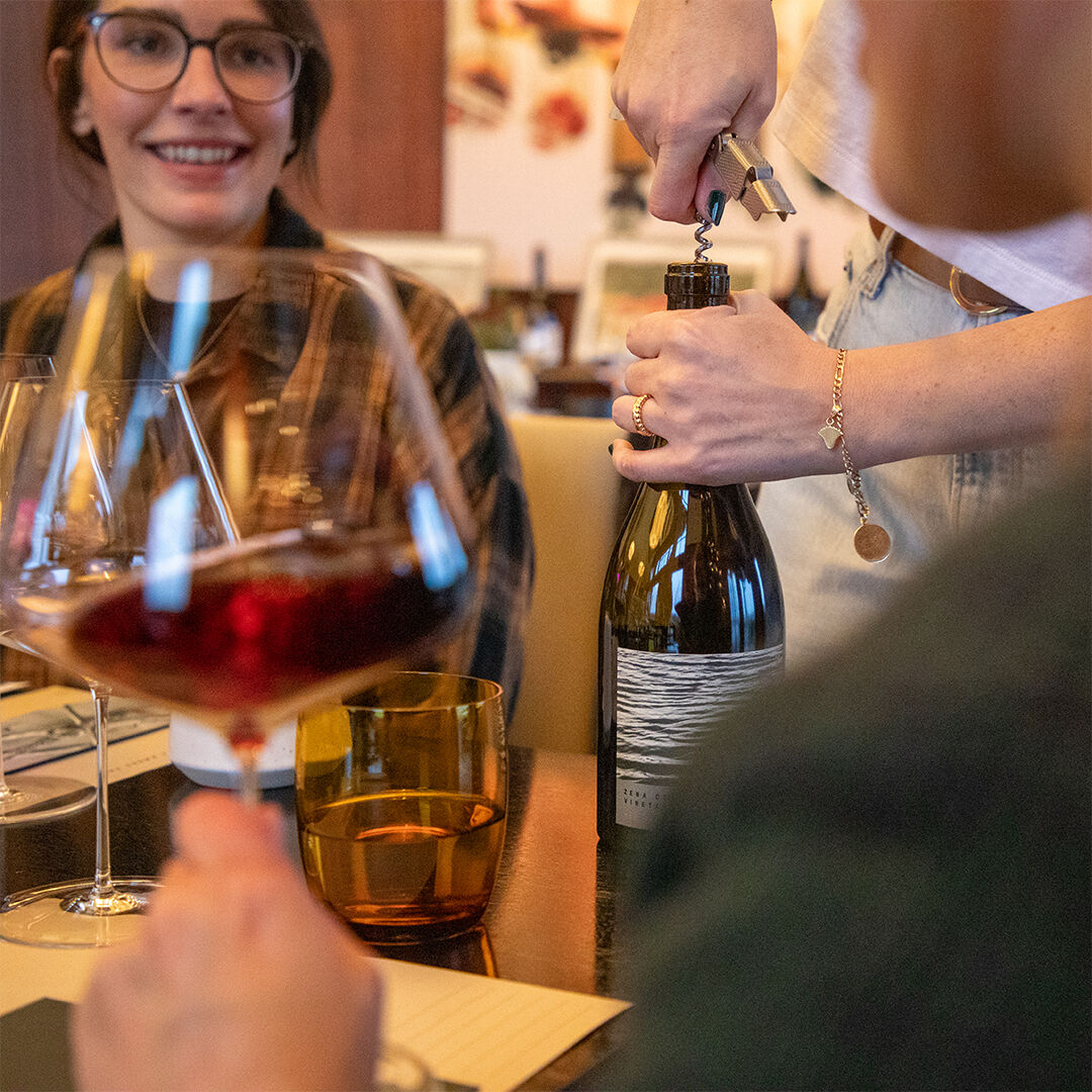 2 women chatting and swirling wine glasses as bottle is being opened by estate host